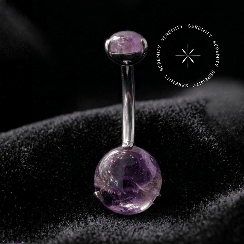 piercing de umbigo titânio repousando suavemente sobre um tecido de cetim branco de alta opulência, destacando o polimento profundo do titânio e os tons roxos da gema natural.