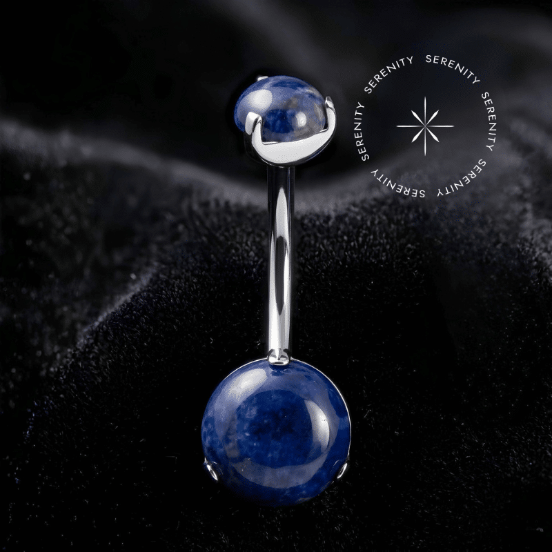 piercing de umbigo titânio repousando suavemente sobre um tecido de cetim branco de alta opulência, destacando o polimento profundo do titânio e os tons azuis da gema natural.