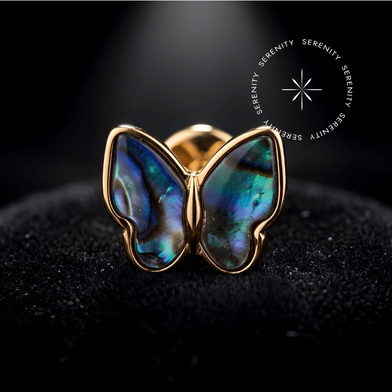 piercing pedra natural titanio repousando suavemente sobre um tecido de cetim preto de alta opulência, destacando sua silhueta de borboleta e o brilho iridescente da gema orgânica abalone.