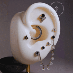 : Piercing titânio compondo um projeto auricular completo repousando suavemente sobre um tecido de cetim branco de alta opulência, evidenciando o labret titânio grau implante, o piercing de helix com correntes e o piercing para conch em argola, destacando o forte contraste entre a estrutura perfeitamente polida, as gemas translúcidas e as pedras negras de lapidação premium.